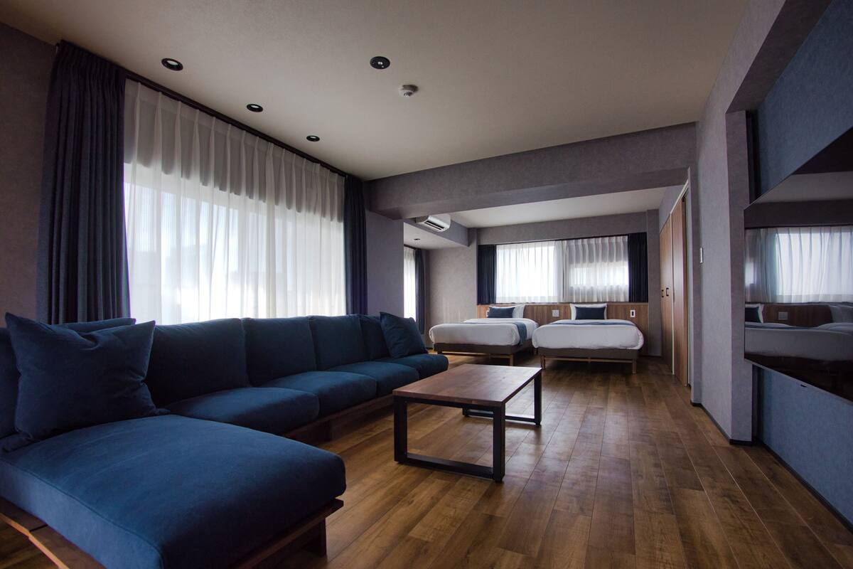 A modern hotel room with a sofa, a small table, two beds, and a large window with curtains.
