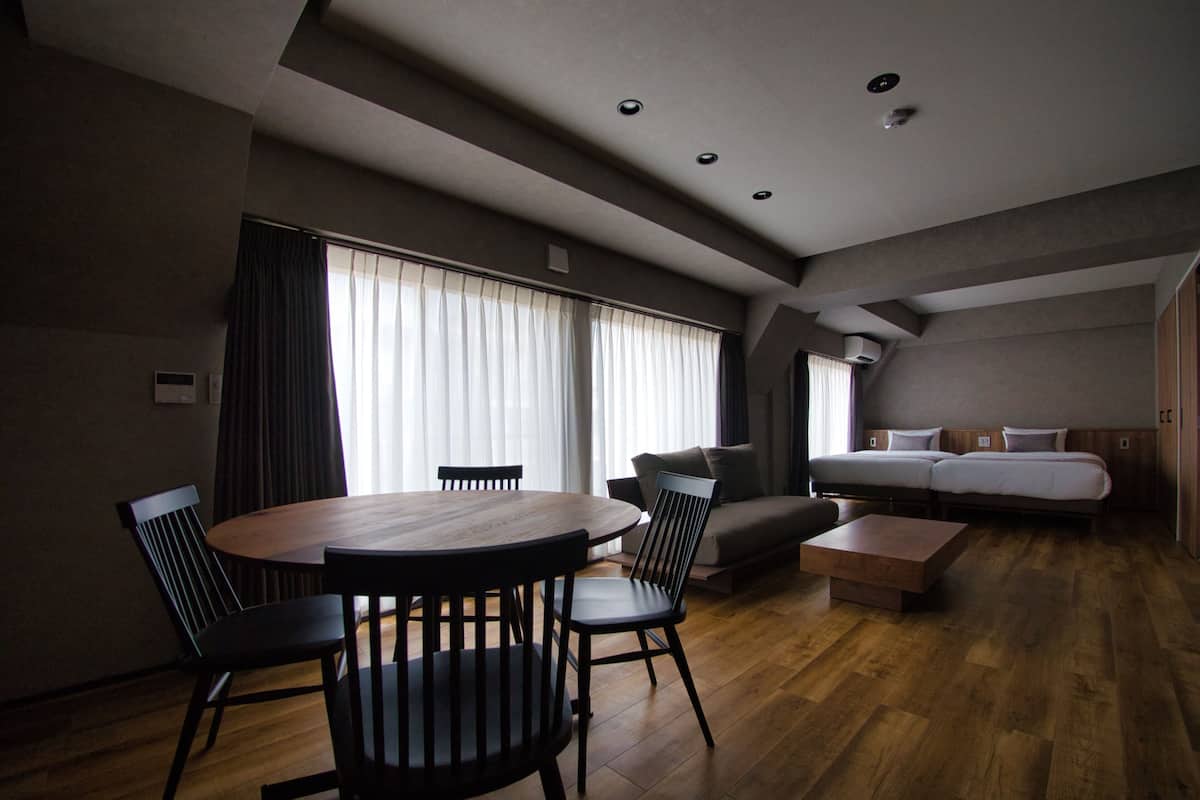 A modern hotel room with a wooden dining table, chairs, a sofa, and a bed.