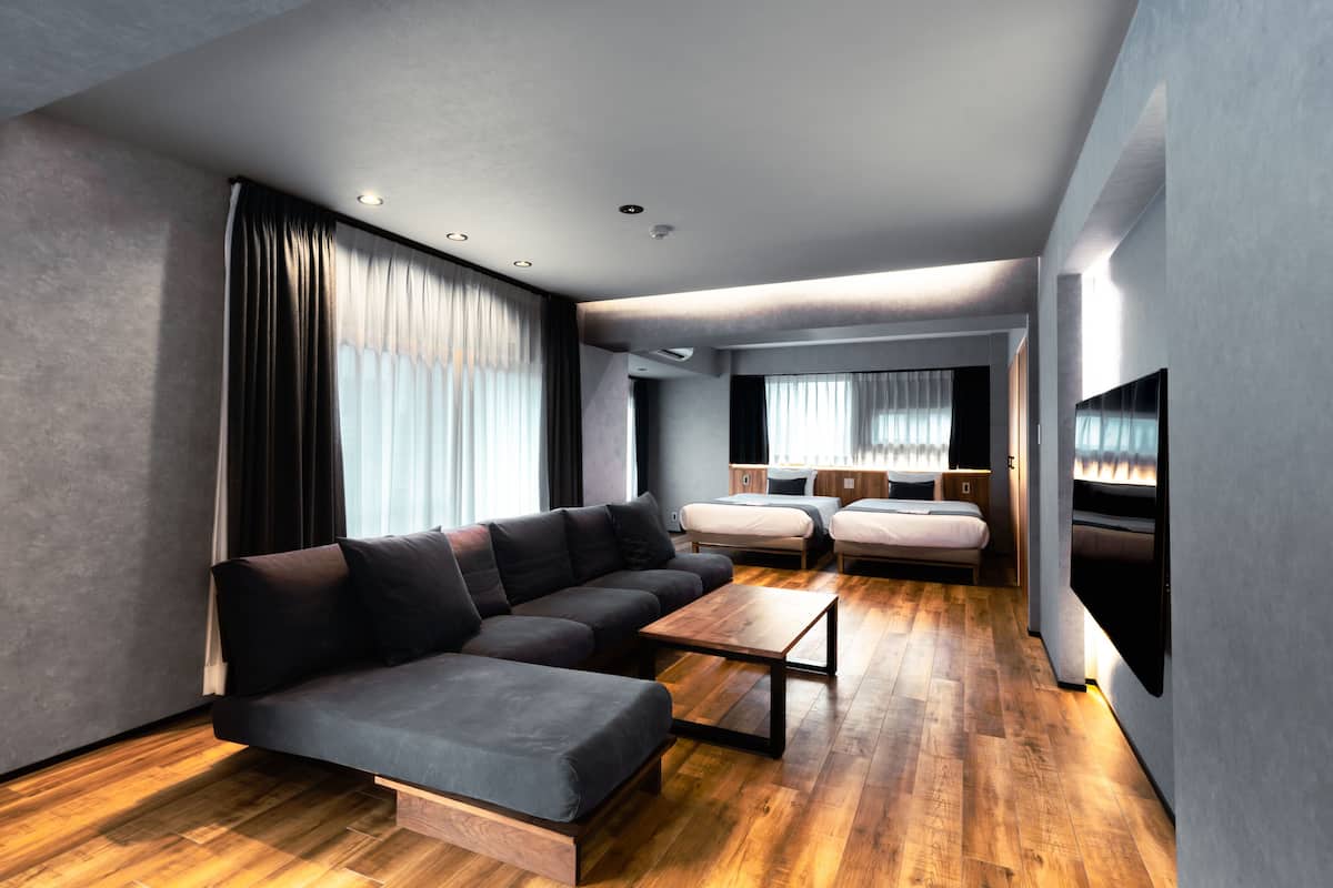 A modern hotel room with a sectional sofa, a small wooden coffee table, a bed, and a flat-screen TV mounted on the wall.