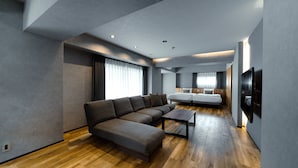 Una sala moderna con un sofá modular, una mesa de centro, un televisor montado en la pared y una zona de dormitorio separada con una cama y una ventana.