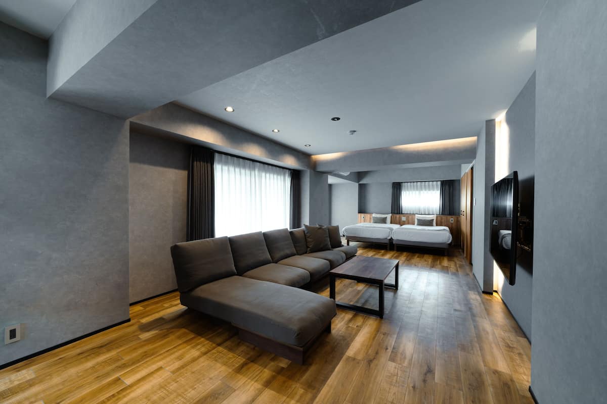 A modern living room with a sectional sofa, a coffee table, a TV mounted on the wall, and a separate bedroom area with a bed and a window.