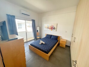 Een moderne hotelkamer met een groot bed, een flatscreen televisie, een schilderij van zeilboten en een balkon met gordijnen.