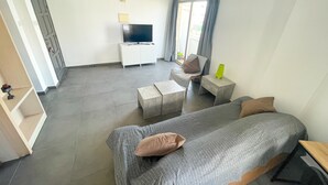 Una habitación de hotel moderna con una cama grande, un televisor sobre un mueble blanco, una zona de estar con una silla y una mesita, y una ventana con cortinas.