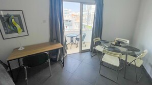 Una habitación pequeña y bien iluminada con un escritorio de madera, una silla, una mesita y un balcón con vistas a edificios.