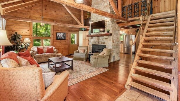 A spacious log cabin interior with a stone fireplace, wooden staircase, and a cozy living area with sofas and a coffee table.