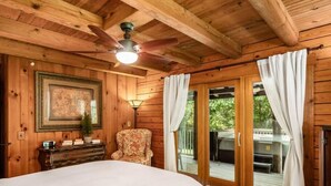 A wooden cabin bedroom with a bed, a chair, a desk, a ceiling fan, and a sliding glass door leading to an outdoor area.