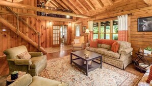 A rustic wooden interior with a high ceiling, a staircase, a sofa, a coffee table, and a patterned rug.