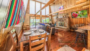 Interior rústico de una cabaña con comedor, chimenea y un ventanal con vistas a la vegetación.