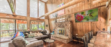 A cozy cabin interior with a stone fireplace, wooden walls, and a large window overlooking a deck.