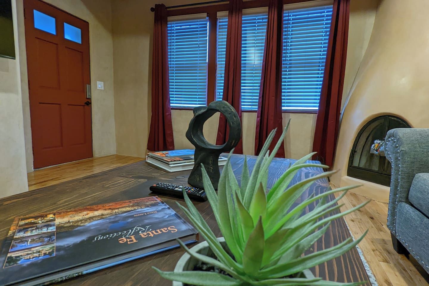 Ein Zimmer mit Holzboden, einer roten Tür, einem Fenster mit Jalousien, einem grauen Sessel, einer Pflanze und einem Buch mit dem Titel 'Santa Fe'.