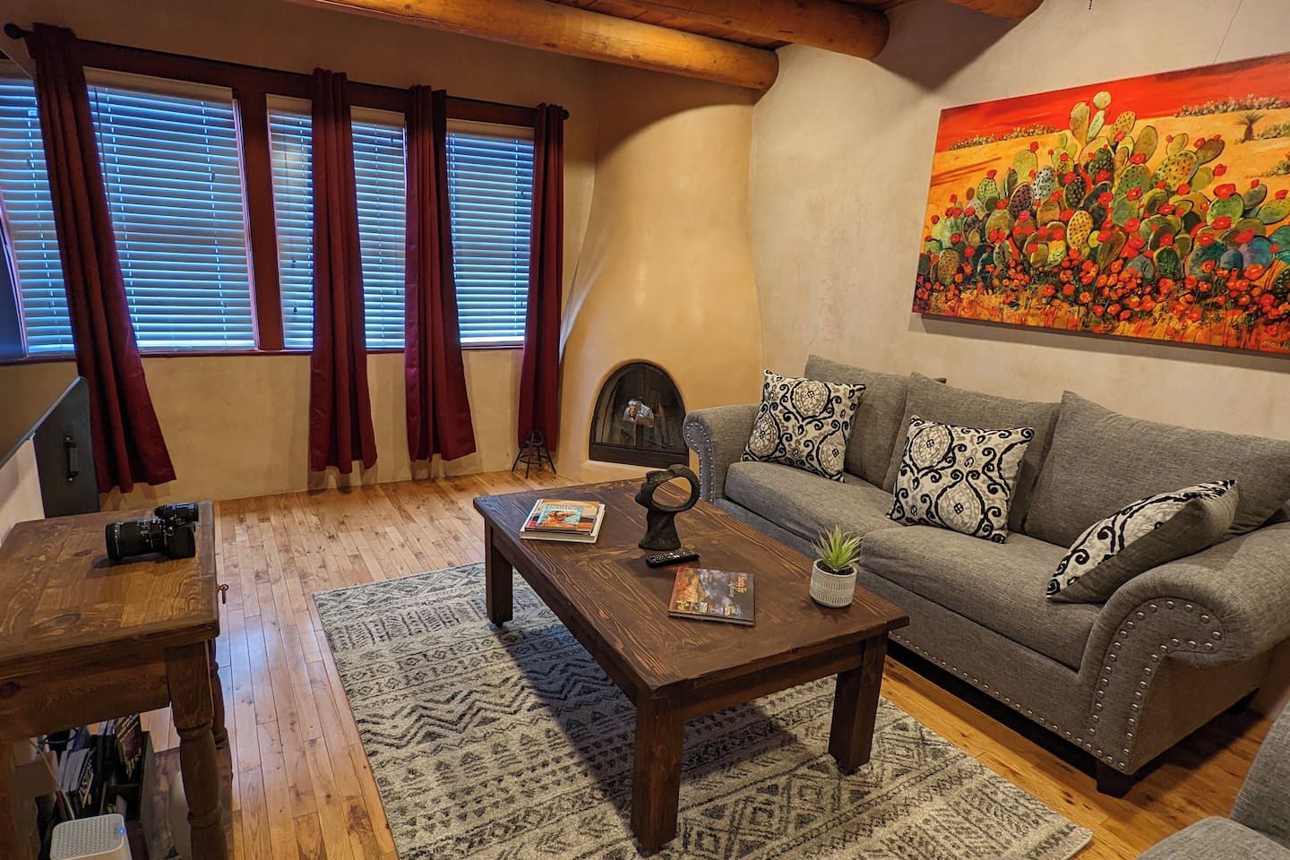 A cozy living room with a wooden coffee table, a sofa with patterned cushions, a fireplace, and a large cactus painting on the wall.