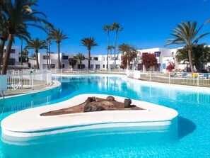 Une piscine agrémentée d’un élément rocheux, entourée de palmiers et de bâtiments blancs.