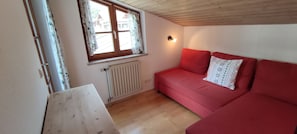 A room with a red sofa, a window with curtains, a radiator, and a wooden ceiling.