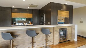 Eine moderne Küche mit dunklen Holzschränken, einer weißen Arbeitsplatte und Edelstahlgeräten. Es gibt drei Barhocker mit Metallbeinen.