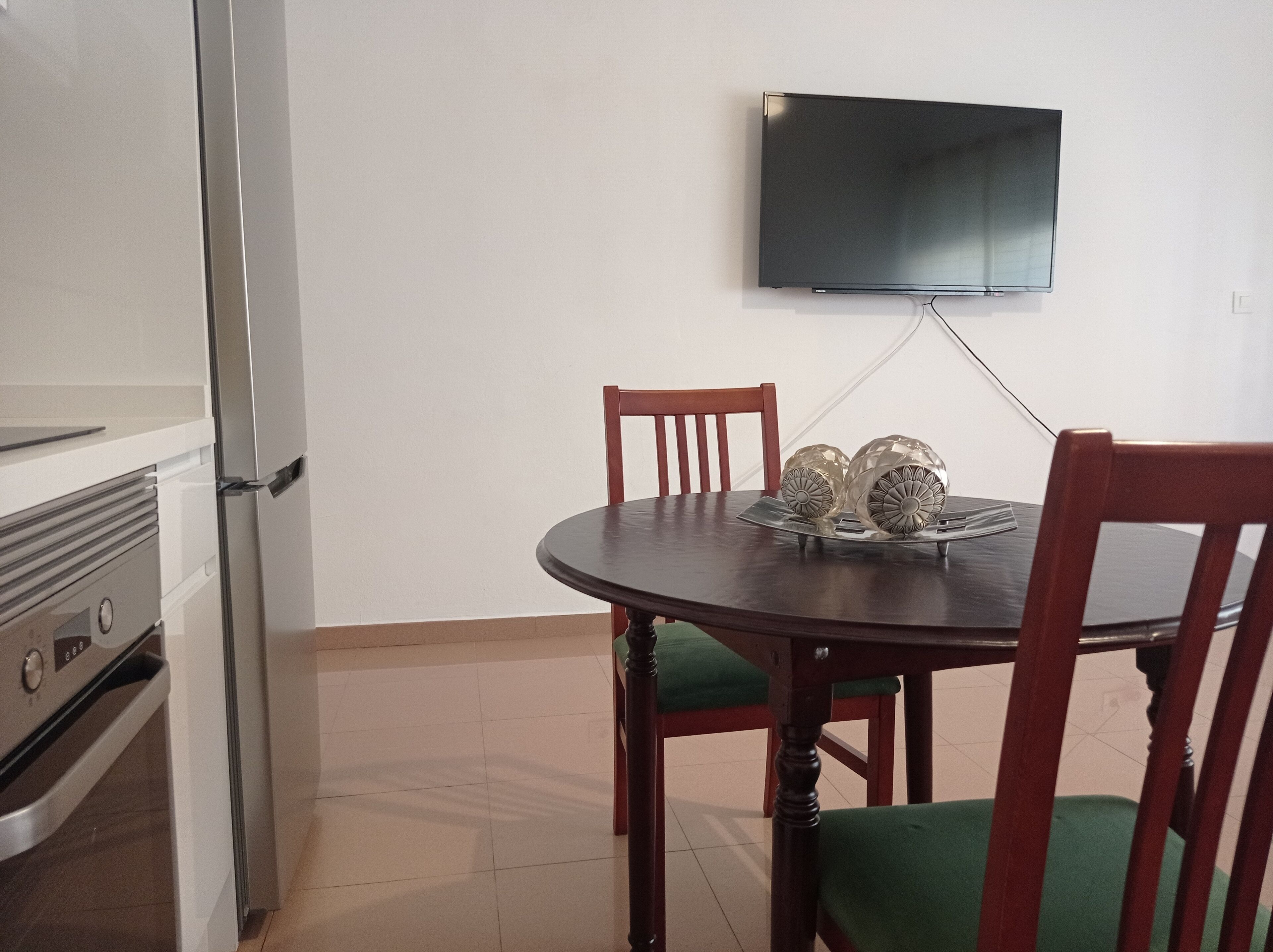 Uma área de refeições com mesa redonda, duas cadeiras, uma televisão de tela plana na parede e uma cozinha com frigorífico e forno.