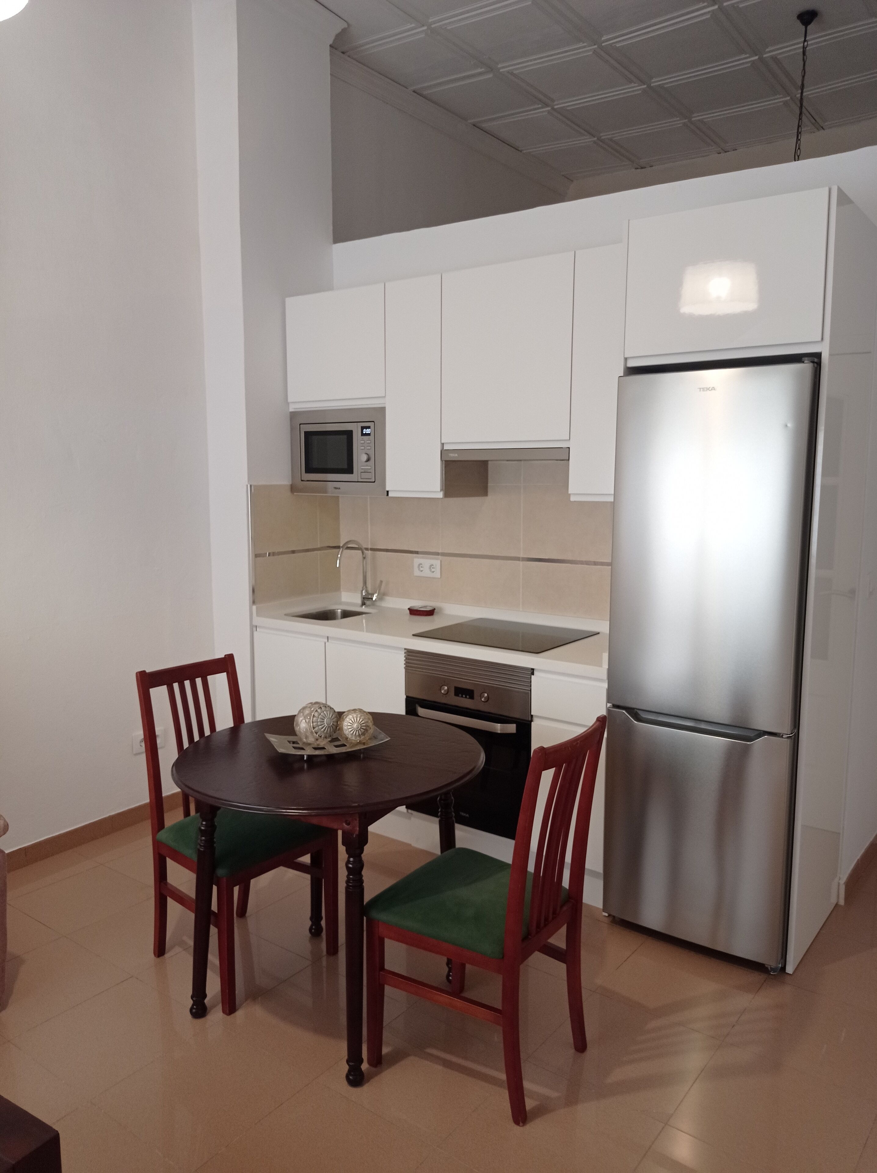 A compact kitchen with white cabinets, a stainless steel refrigerator, a microwave, and a dishwasher. There is a wooden dining table with two chairs.