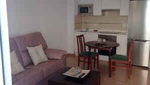 Un salon moderne avec un canapé, une table basse et une table à manger avec des chaises. On aperçoit, au fond, une cuisine avec des meubles blancs et des appareils électroménagers.