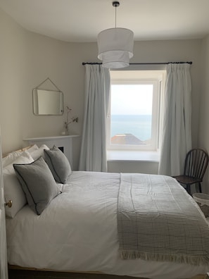 Een slaapkamer met een groot bed, een raam met gordijnen, een hanglamp, een stoel en een tafeltje met een vaas.