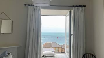 Una camera da letto con un letto, una sedia, una finestra con tende e una lampada a sospensione, con vista sul mare e sugli edifici.