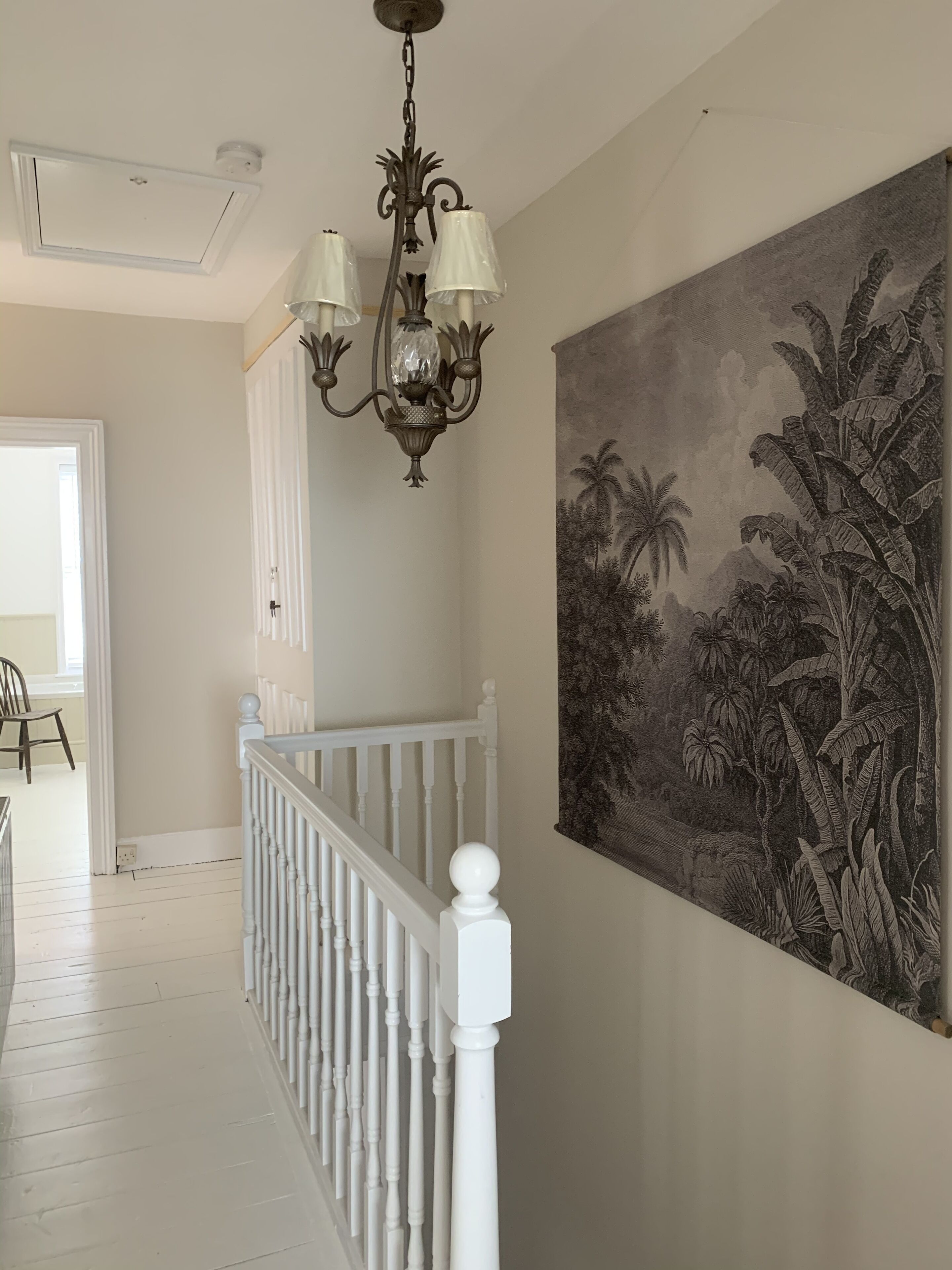 A hallway with a white railing, a chandelier, and a large tropical-themed painting on the wall.