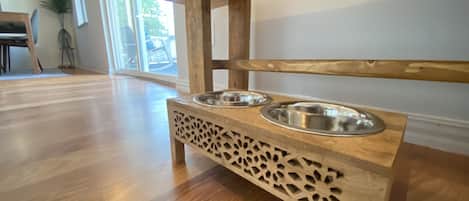 A wooden pet feeding station with two metal bowls, located in a room with wooden flooring and a large window.