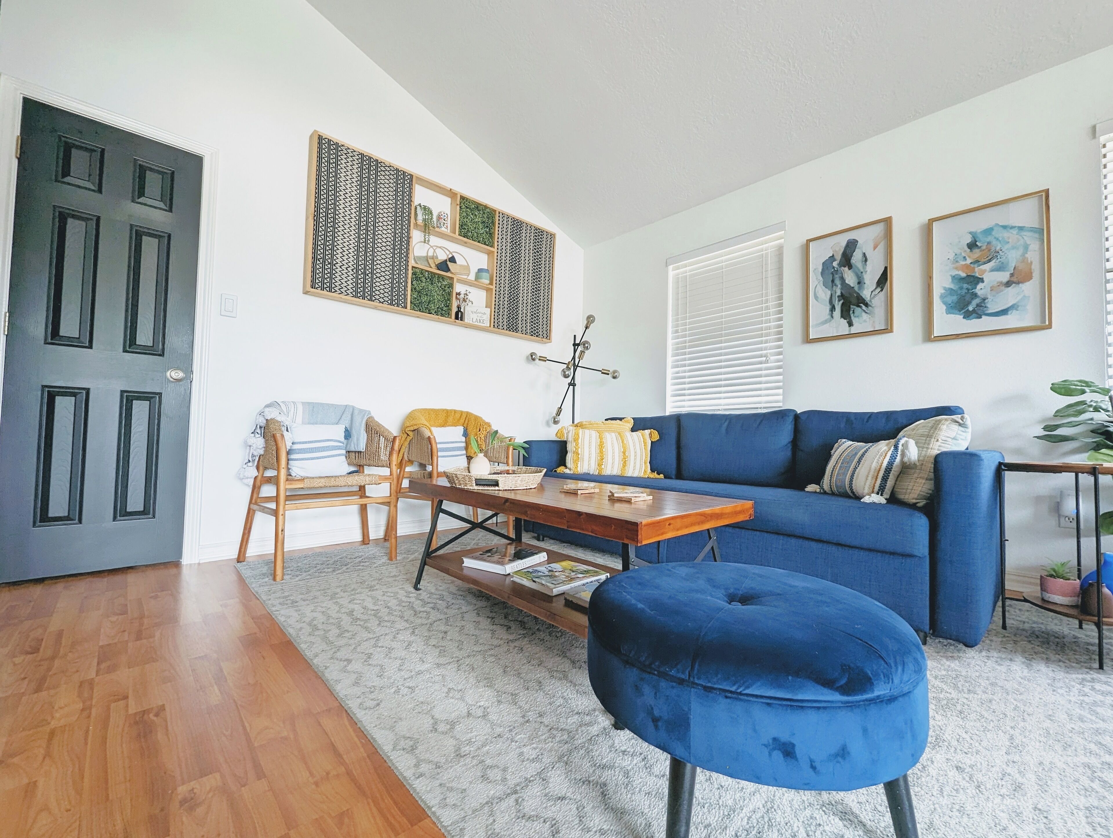 A living room with a blue sofa, a wooden coffee table, and a blue ottoman. There is a door, a wall-mounted shelf with decorative items, and a window with blinds.