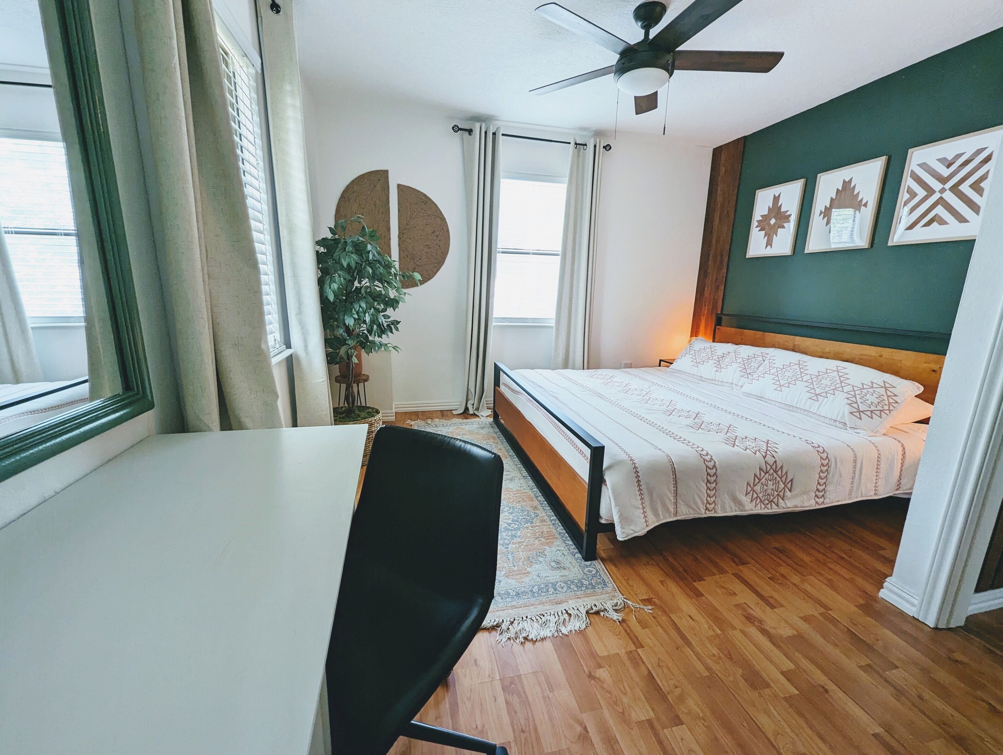 Ein Schlafzimmer mit Bett, Schreibtisch, Stuhl, Deckenventilator, einem Fenster mit Vorhängen, einer Topfpflanze und Wandbildern.