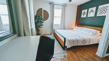 Ein Schlafzimmer mit Bett, Schreibtisch, Stuhl, Deckenventilator, einem Fenster mit Vorhängen, einer Topfpflanze und Wandbildern.
