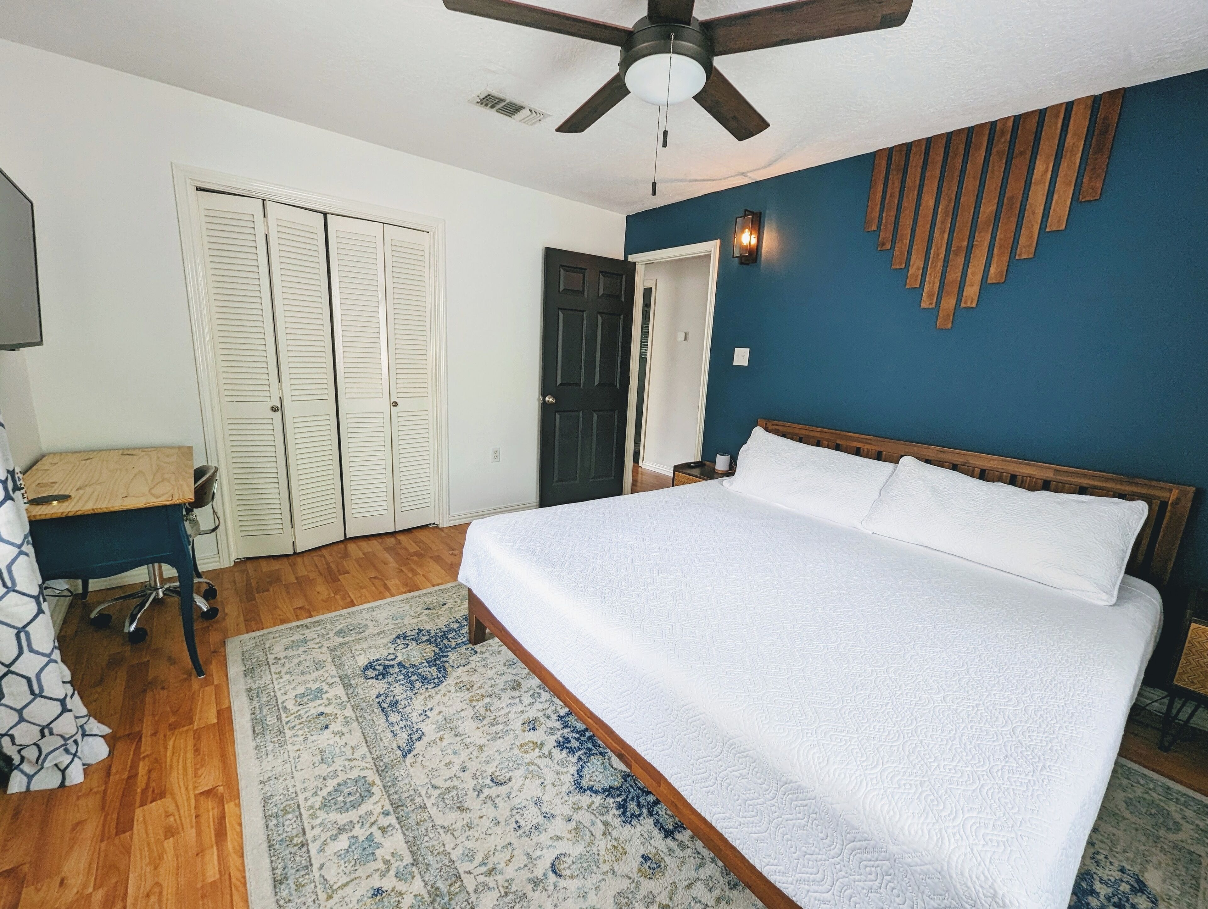 A bedroom with a bed, a desk, a chair, a ceiling fan, and a door.