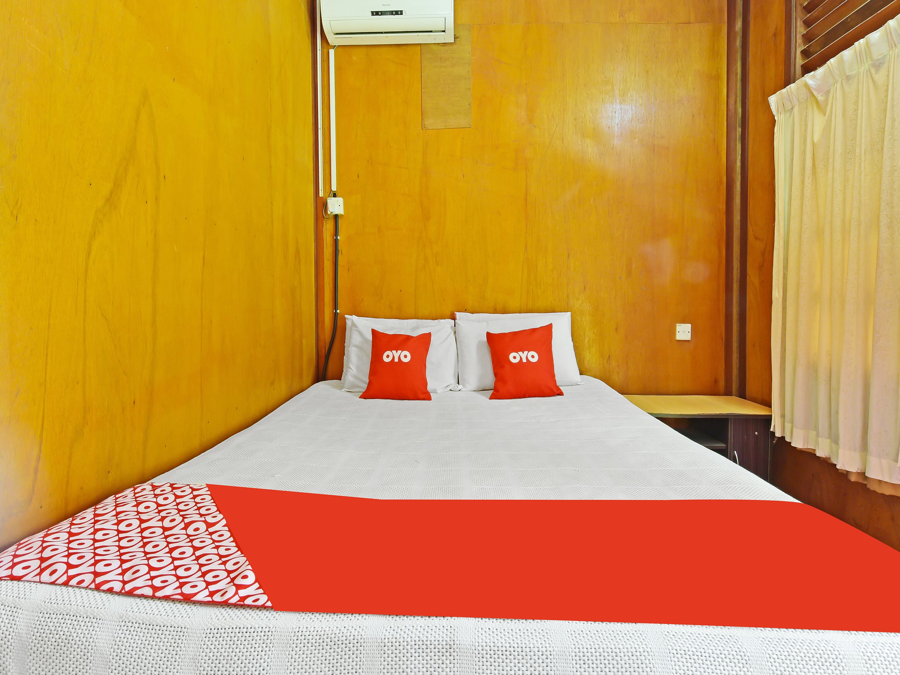 Une chambre d’hôtel avec un mur en bois, un lit avec une literie blanche et deux oreillers rouges, une couverture rouge et blanche, et une fenêtre avec des rideaux.