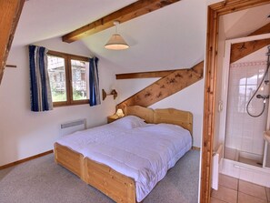A bedroom with a wooden bed, a window with curtains, a wall-mounted lamp, and a shower area.