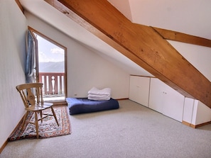 A small, sloped room with a wooden ceiling, a single bed with a blue mattress, a wooden chair, a small rug, and a view of mountains through a window.