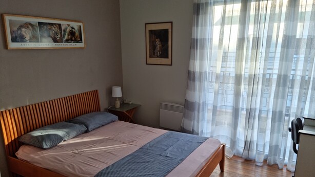 A bedroom with a wooden bed, a nightstand with a lamp, a framed picture on the wall, and a window with sheer curtains. - Stadion apt 2 Near Athens city center (Athina)