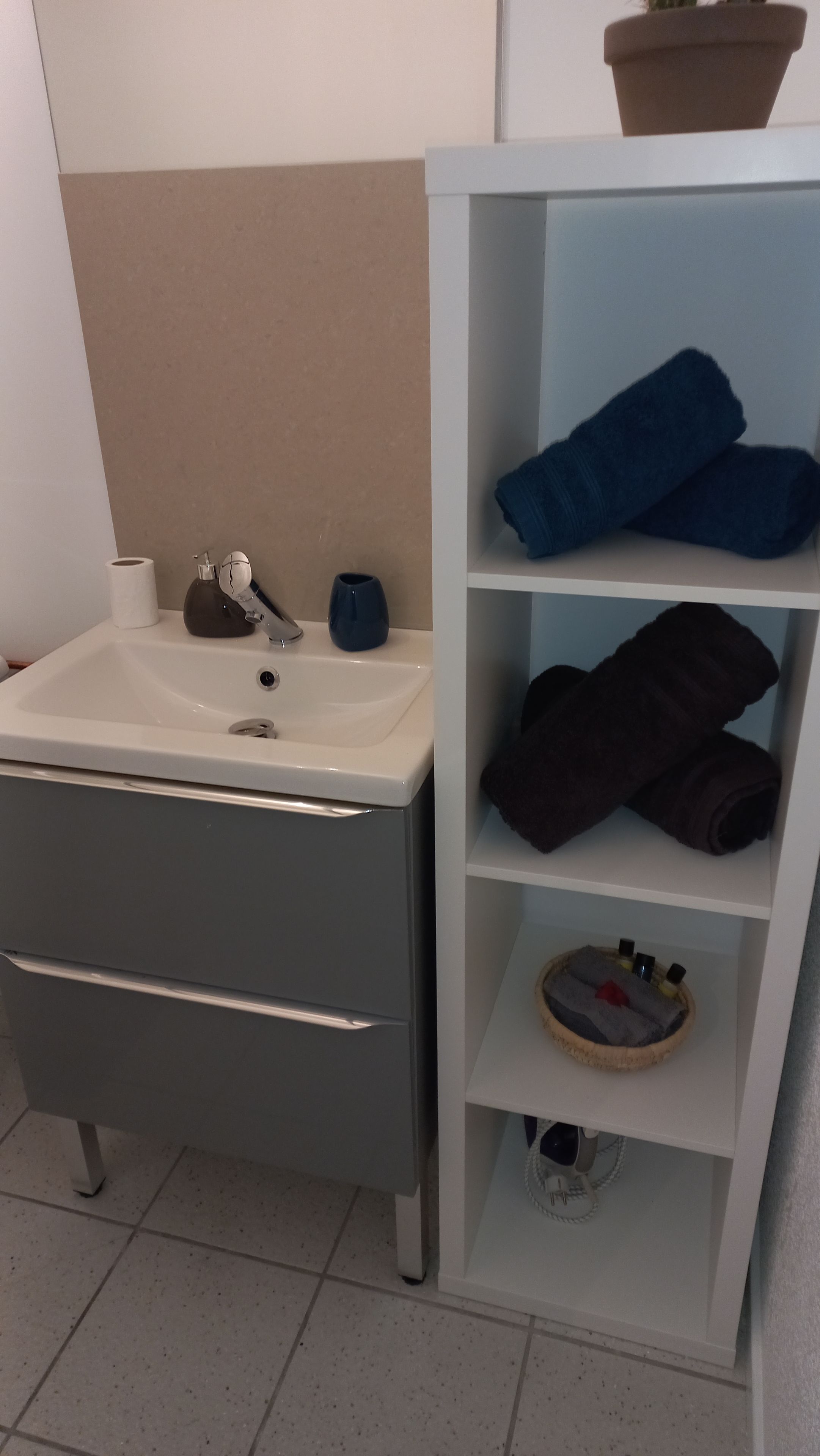 A bathroom with a white sink, a grey cabinet, and a shelf with towels and toiletries.
