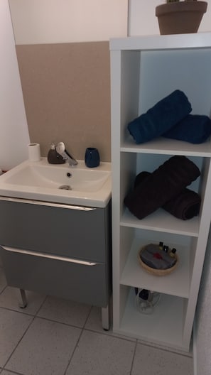 Ett badrum med ett vitt handfat, ett grått skåp och en hylla med handdukar och toalettartiklar.