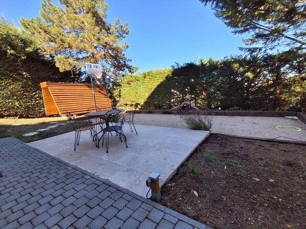 A patio with a table and chairs, surrounded by a garden and a wooden structure.