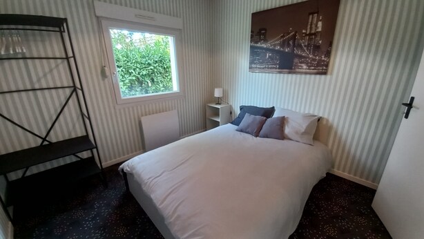 Una habitació d'hotel amb un llit, una finestra, un radiador, una taula i un quadre d'un paisatge urbà.