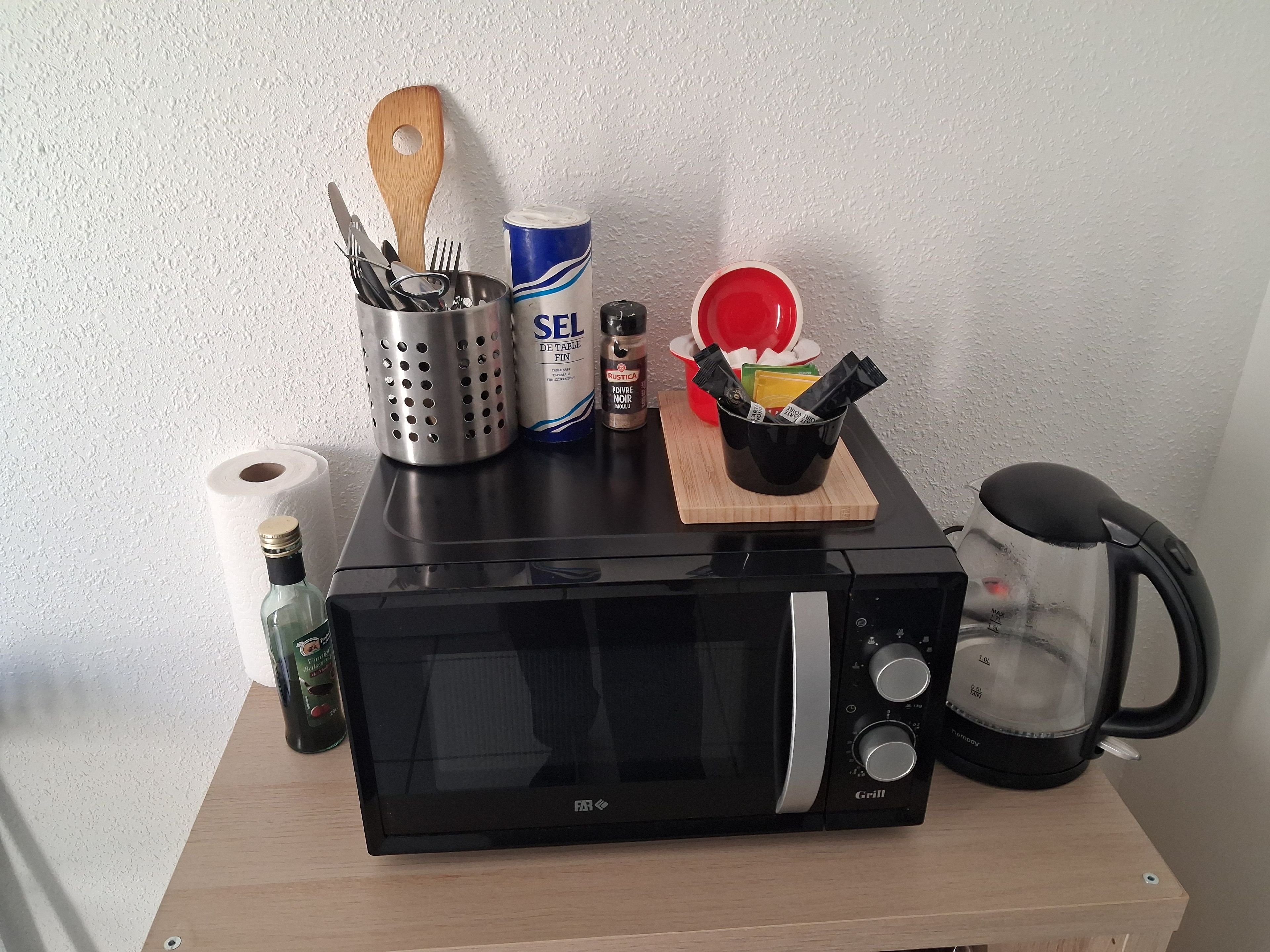 Een magnetron op een houten tafel, samen met een koffiezetapparaat, een rol keukenpapier, een fles, een bak met bestek en een blik kookolie.