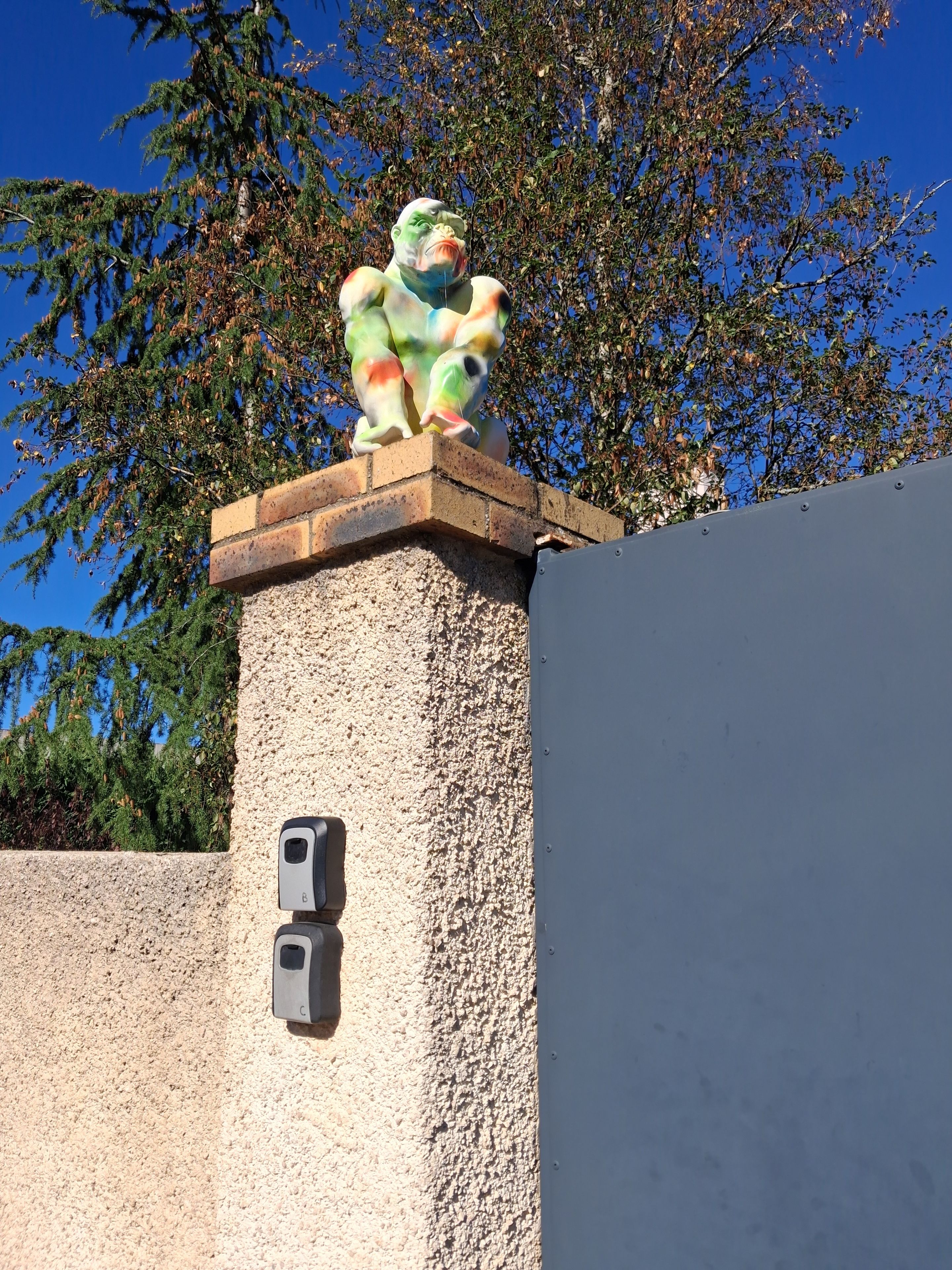 Une statue posée sur une corniche en briques, devant un mur texturé et une boîte en métal.