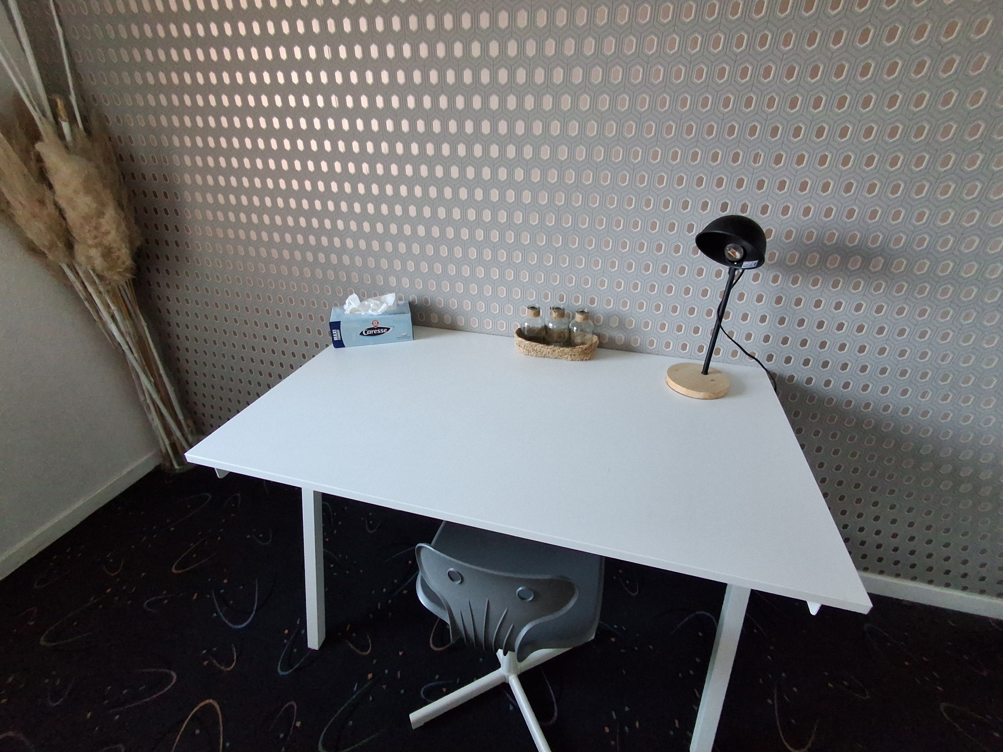 A white desk with a lamp, a tissue box, and a basket with jars against a perforated wall.