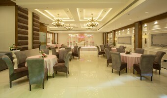 A spacious hotel dining area with multiple round tables and chairs, elegant chandeliers, and a stage with floral decorations.