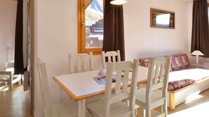 Une pièce comprenant un lit, une table et des chaises. Il y a une fenêtre avec des rideaux, un tableau au mur et une lampe.
