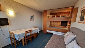 Ein Etagenbett mit Holzrahmen, die untere Liege mit einer Matratze. Rechts daneben ein Einzelbett mit Matratze und Kissen. Vor dem Etagenbett steht ein Tisch mit weißem Tischläufer und zwei Stühle. Ein Fernseher ist an der linken Wand befestigt.