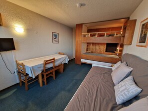 A bunk bed with a wooden frame and a lower bunk with a mattress. There is a single bed with a mattress and pillows on the right. A table with a white tablecloth and two chairs are positioned in front of the bunk bed. A television is mounted on the wall to the left.