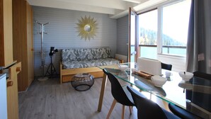 Una sala de estar moderna con un sofá, una mesa de vidrio y sillas. Una ventana grande ofrece vistas del exterior.