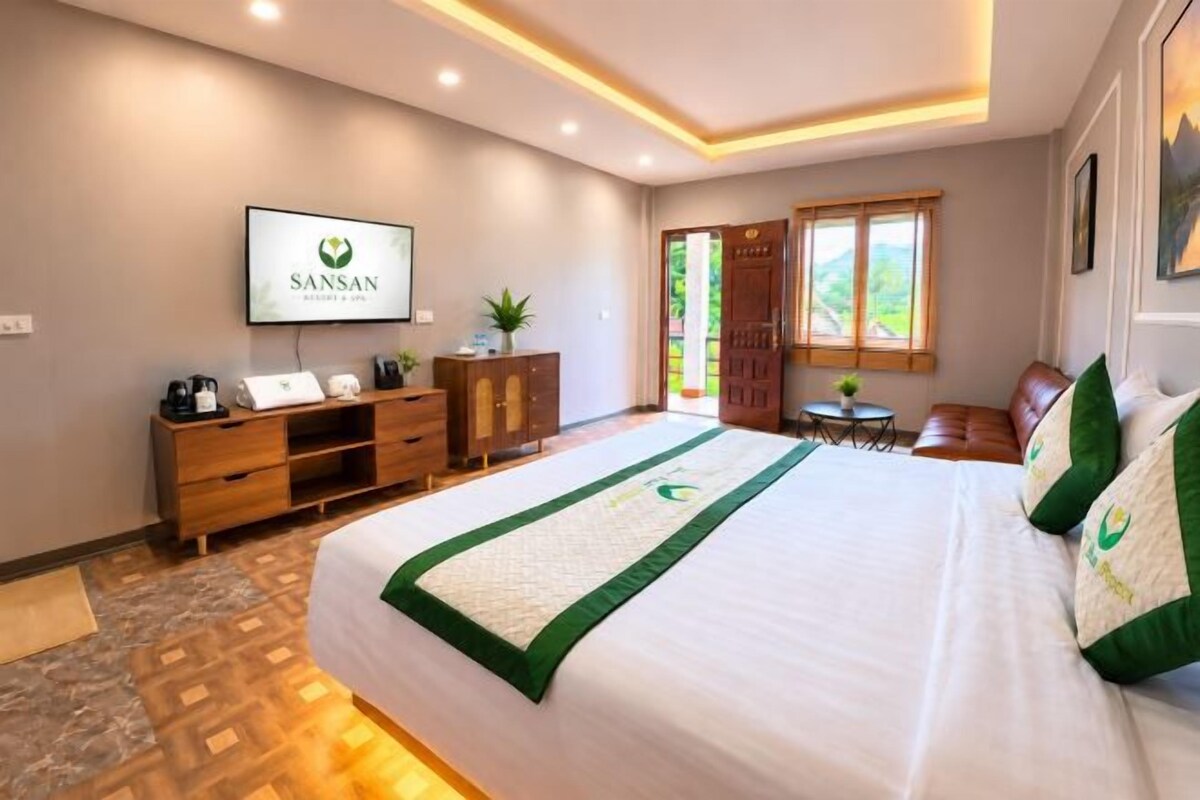 A hotel room with a bed, a television, a desk with a plant, and a window with a view of greenery.