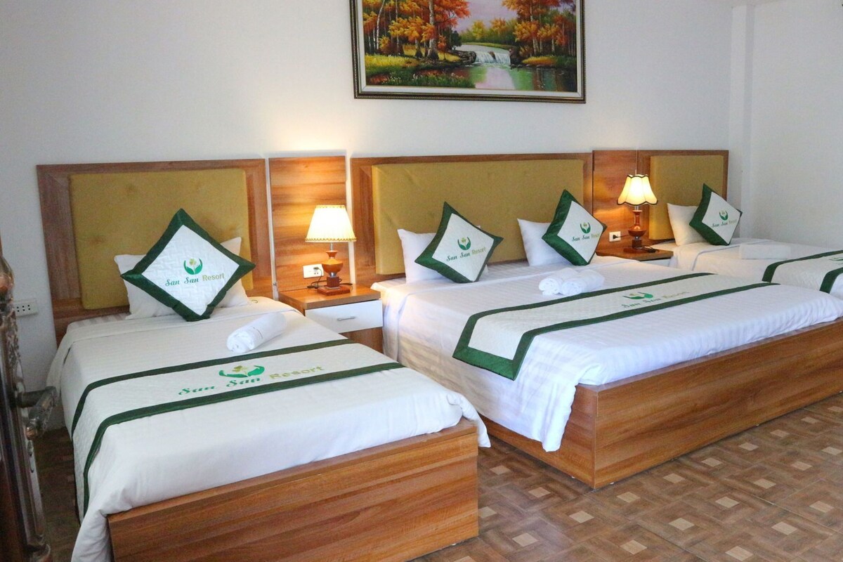 A hotel room with four beds, each with a green and white bedspread and matching pillows. There are bedside lamps and a painting of a landscape on the wall.