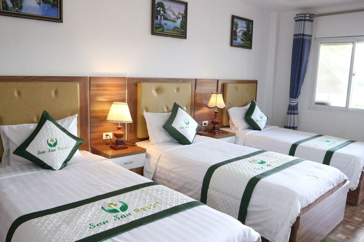 A hotel room with four beds, each with a headboard and a pillow with the hotel's logo. There are bedside lamps, a window with curtains, and framed pictures on the wall.