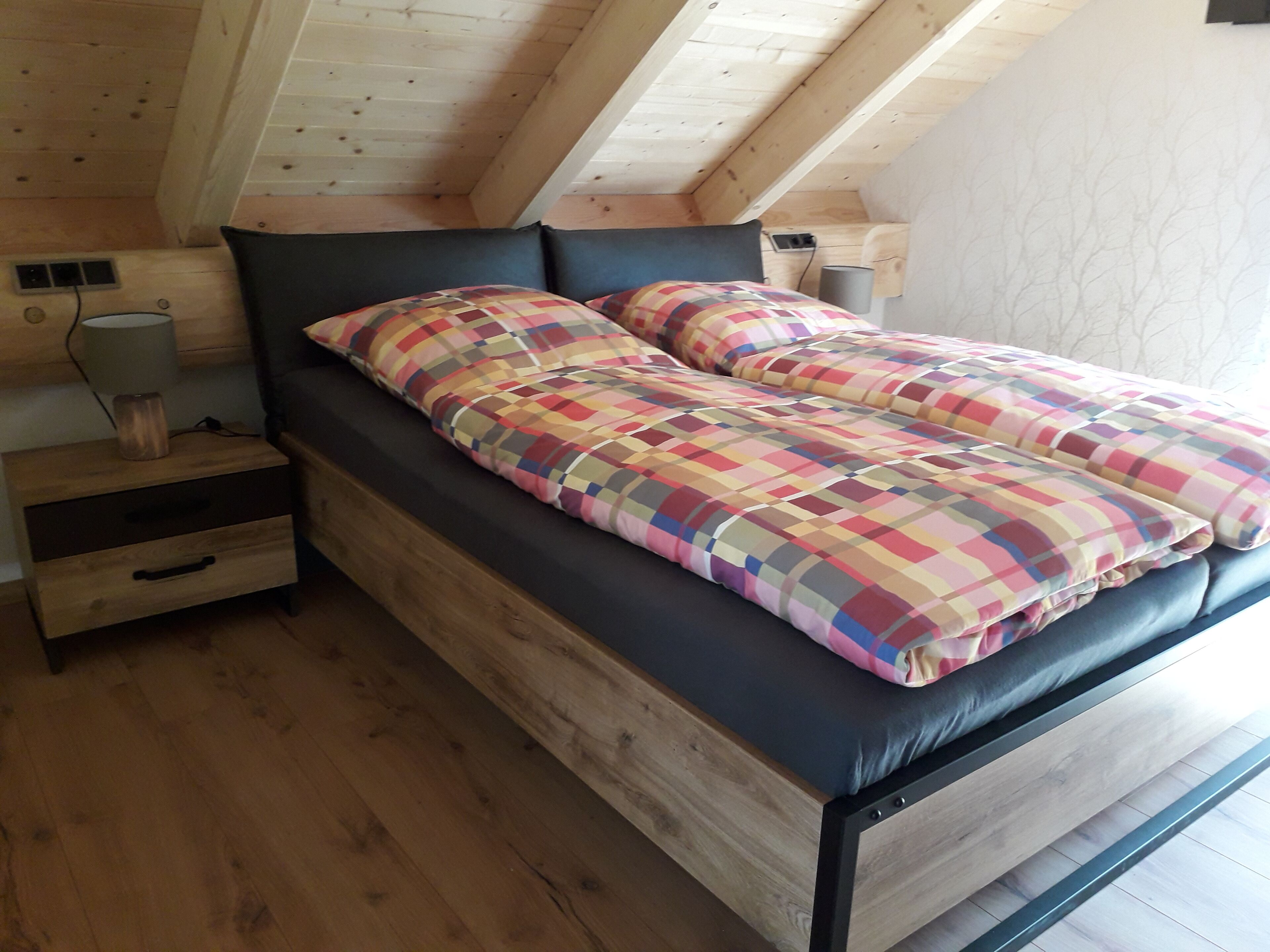 A wooden bed with a black frame and colorful checkered bedding in a room with wooden floors and sloped ceilings.