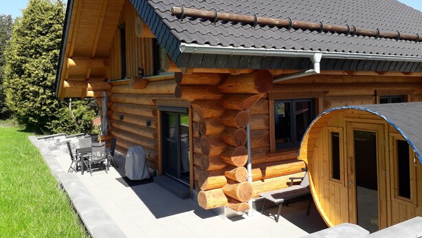 A log cabin with a tiled roof, a covered patio with outdoor furniture, and a large wooden door. - Kanadisches Naturstamm-blockhaus (Linden)